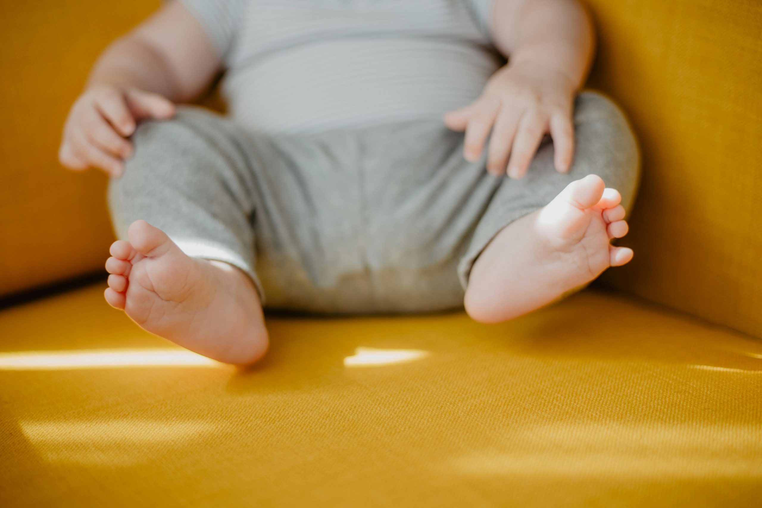 Pieds de bébé en gros plan. Le bébé est assis sur un fauteuil jaune. Les doigts de pied sont en éventail. On ne voit que le bas du corps.