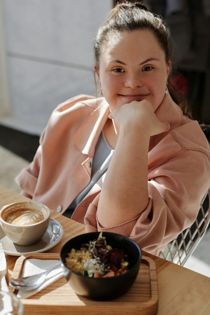Jeune femme porteuse de trisomie 21 souriant, assise à une table, une main devant sa bouche.